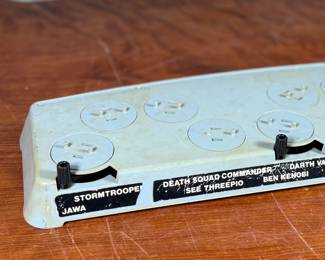 Vintage 1978 Star Wars action figure display stand by GMFGI. Marked ‘321E 001’ with a copyright of ‘G.M.F.G.I. – 1978’. Features slots marked with character names including Stormtrooper, Darth Vader, Luke Skywalker, and Princess Leia. Made of gray plastic with attached gear mechanisms on the back and visible character labels on the front. Some wear consistent with age.