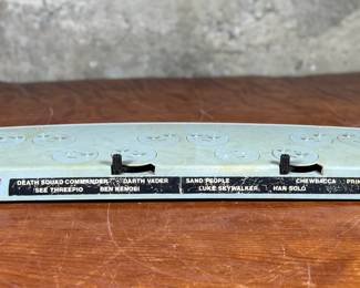 Vintage 1978 Star Wars action figure display stand by GMFGI. Marked ‘321E 001’ with a copyright of ‘G.M.F.G.I. – 1978’. Features slots marked with character names including Stormtrooper, Darth Vader, Luke Skywalker, and Princess Leia. Made of gray plastic with attached gear mechanisms on the back and visible character labels on the front. Some wear consistent with age.