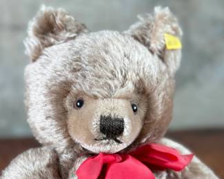 Steiff mohair teddy bear featuring a soft brown mohair exterior and traditional button-in-ear yellow tag. Adorned with a red bow around the neck, this bear’s label indicates it is made of 53% wool and 47% cotton. Includes an “Original Teddybar” tag and is in great collectible condition.