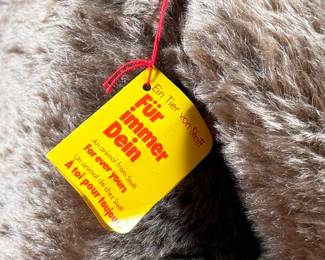 Steiff mohair teddy bear featuring a soft brown mohair exterior and traditional button-in-ear yellow tag. Adorned with a red bow around the neck, this bear’s label indicates it is made of 53% wool and 47% cotton. Includes an “Original Teddybar” tag and is in great collectible condition.