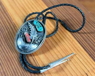Silver bolo tie featuring a decorative oval slide adorned with turquoise and coral cabochons set in an intricate feather motif. The tie features a black braided leather cord with silver-tone tips and is marked ‘Bennett Pat Pend’ on the back. Shows slight age-related wear on metal surfaces consistent with use.