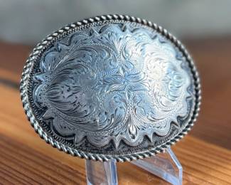 Sterling silver belt buckle crafted by Kustom Kraft featuring an intricate engraved pattern with foliate and scroll motifs. The oval-shaped buckle is bordered with a twisted rope-style edge. The back is stamped with ‘STERLING’ and the Kustom Kraft hallmark. Measures, 3” H x 4” W with a total approx. weight of 87.00 grams.
