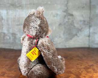 Steiff mohair teddy bear featuring a soft brown mohair exterior and traditional button-in-ear yellow tag. Adorned with a red bow around the neck, this bear’s label indicates it is made of 53% wool and 47% cotton. Includes an “Original Teddybar” tag and is in great collectible condition.