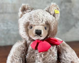 Steiff mohair teddy bear featuring a soft brown mohair exterior and traditional button-in-ear yellow tag. Adorned with a red bow around the neck, this bear’s label indicates it is made of 53% wool and 47% cotton. Includes an “Original Teddybar” tag and is in great collectible condition.