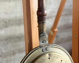 Brass bed warmer with floral etching and decorative perforations on the lid. The piece features a wooden turned handle with a rich patina and a hinged lid. The brass showcases signs of use with wear and age-related patina. The perforations were traditionally used for dispersing heat by placing warm materials inside the bed warmer. Suitable for collectors or historical display.