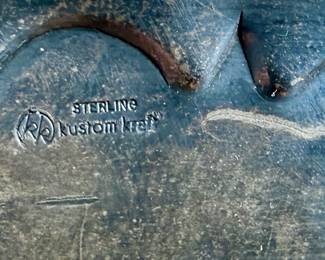 Sterling silver belt buckle crafted by Kustom Kraft featuring an intricate engraved pattern with foliate and scroll motifs. The oval-shaped buckle is bordered with a twisted rope-style edge. The back is stamped with ‘STERLING’ and the Kustom Kraft hallmark. Measures, 3” H x 4” W with a total approx. weight of 87.00 grams.