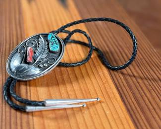 Silver bolo tie featuring a decorative oval slide adorned with turquoise and coral cabochons set in an intricate feather motif. The tie features a black braided leather cord with silver-tone tips and is marked ‘Bennett Pat Pend’ on the back. Shows slight age-related wear on metal surfaces consistent with use.