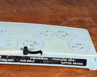 Vintage 1978 Star Wars action figure display stand by GMFGI. Marked ‘321E 001’ with a copyright of ‘G.M.F.G.I. – 1978’. Features slots marked with character names including Stormtrooper, Darth Vader, Luke Skywalker, and Princess Leia. Made of gray plastic with attached gear mechanisms on the back and visible character labels on the front. Some wear consistent with age.