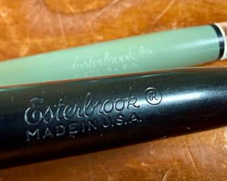 Esterbrook dip pens featuring metal nibs marked ‘Esterbrook 9556’ with black and green handles. Each pen is paired with a ceramic holder in complementing colors: green and blue-gray. Both pens are marked ‘Esterbrook Made in U.S.A.’ on the handles, denoting their authenticity. Well-preserved and functional writing tools, ideal for collectors or calligraphy enthusiasts.