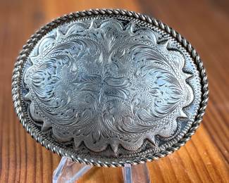 Sterling silver belt buckle crafted by Kustom Kraft featuring an intricate engraved pattern with foliate and scroll motifs. The oval-shaped buckle is bordered with a twisted rope-style edge. The back is stamped with ‘STERLING’ and the Kustom Kraft hallmark. Measures, 3” H x 4” W with a total approx. weight of 87.00 grams.