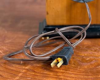 Vintage Chronomaster electric clock featuring a wood housing with black base and decorative brass-tone accents. The clock measures 9” in height and includes an electrical cord for operation. The back panel displays the manufacturer’s details, marked ‘Made in U.S.A.’ and specifications for 115V-60 cycle-2W. Tested and in working condition.