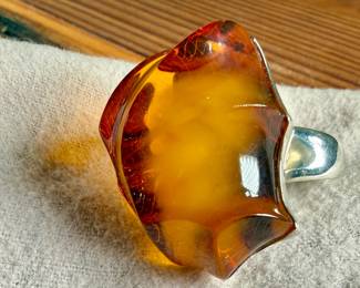Sterling silver ring featuring a large, polished amber cabochon centerpiece with organic and irregular contours. The amber showcases rich honey tones with natural inclusions visible within. The mounting is made from solid sterling silver and complements the amber’s unique shape. Coin shown for size reference. Comes in a size 9 with a total approx. weight of 27.21 grams.