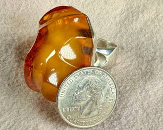 Sterling silver ring featuring a large, polished amber cabochon centerpiece with organic and irregular contours. The amber showcases rich honey tones with natural inclusions visible within. The mounting is made from solid sterling silver and complements the amber’s unique shape. Coin shown for size reference. Comes in a size 9 with a total approx. weight of 27.21 grams.
