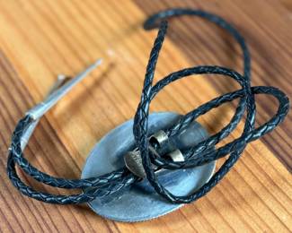 Silver bolo tie featuring a decorative oval slide adorned with turquoise and coral cabochons set in an intricate feather motif. The tie features a black braided leather cord with silver-tone tips and is marked ‘Bennett Pat Pend’ on the back. Shows slight age-related wear on metal surfaces consistent with use.