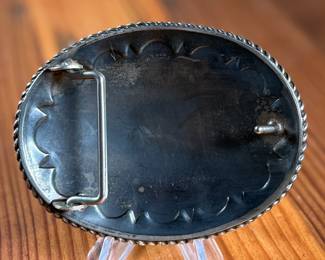 Sterling silver belt buckle crafted by Kustom Kraft featuring an intricate engraved pattern with foliate and scroll motifs. The oval-shaped buckle is bordered with a twisted rope-style edge. The back is stamped with ‘STERLING’ and the Kustom Kraft hallmark. Measures, 3” H x 4” W with a total approx. weight of 87.00 grams.