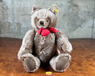 Steiff mohair teddy bear featuring a soft brown mohair exterior and traditional button-in-ear yellow tag. Adorned with a red bow around the neck, this bear’s label indicates it is made of 53% wool and 47% cotton. Includes an “Original Teddybar” tag and is in great collectible condition.