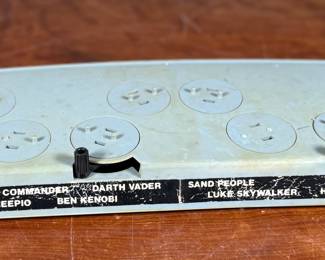 Vintage 1978 Star Wars action figure display stand by GMFGI. Marked ‘321E 001’ with a copyright of ‘G.M.F.G.I. – 1978’. Features slots marked with character names including Stormtrooper, Darth Vader, Luke Skywalker, and Princess Leia. Made of gray plastic with attached gear mechanisms on the back and visible character labels on the front. Some wear consistent with age.