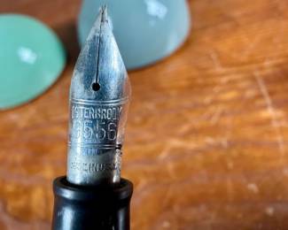 Esterbrook dip pens featuring metal nibs marked ‘Esterbrook 9556’ with black and green handles. Each pen is paired with a ceramic holder in complementing colors: green and blue-gray. Both pens are marked ‘Esterbrook Made in U.S.A.’ on the handles, denoting their authenticity. Well-preserved and functional writing tools, ideal for collectors or calligraphy enthusiasts.