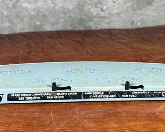 Vintage 1978 Star Wars action figure display stand by GMFGI. Marked ‘321E 001’ with a copyright of ‘G.M.F.G.I. – 1978’. Features slots marked with character names including Stormtrooper, Darth Vader, Luke Skywalker, and Princess Leia. Made of gray plastic with attached gear mechanisms on the back and visible character labels on the front. Some wear consistent with age.