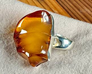 Sterling silver ring featuring a large, polished amber cabochon centerpiece with organic and irregular contours. The amber showcases rich honey tones with natural inclusions visible within. The mounting is made from solid sterling silver and complements the amber’s unique shape. Coin shown for size reference. Comes in a size 9 with a total approx. weight of 27.21 grams.