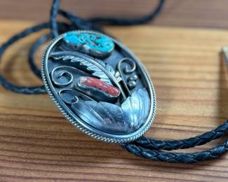 Silver bolo tie featuring a decorative oval slide adorned with turquoise and coral cabochons set in an intricate feather motif. The tie features a black braided leather cord with silver-tone tips and is marked ‘Bennett Pat Pend’ on the back. Shows slight age-related wear on metal surfaces consistent with use.
