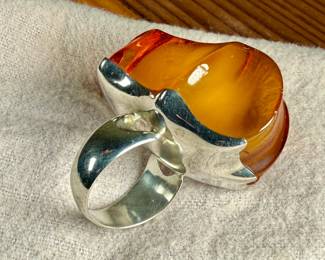 Sterling silver ring featuring a large, polished amber cabochon centerpiece with organic and irregular contours. The amber showcases rich honey tones with natural inclusions visible within. The mounting is made from solid sterling silver and complements the amber’s unique shape. Coin shown for size reference. Comes in a size 9 with a total approx. weight of 27.21 grams.