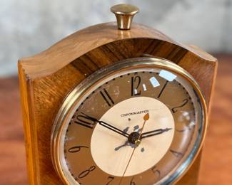 Vintage Chronomaster electric clock featuring a wood housing with black base and decorative brass-tone accents. The clock measures 9” in height and includes an electrical cord for operation. The back panel displays the manufacturer’s details, marked ‘Made in U.S.A.’ and specifications for 115V-60 cycle-2W. Tested and in working condition.