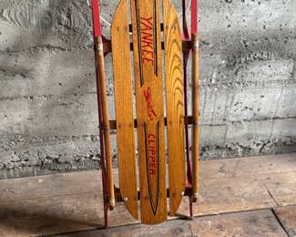 Wooden Flexible Flyer Yankee Clipper sled featuring steel runners painted in red and a wooden deck with printed branding. The sled includes a wooden crossbar for steering and a rope control. Signs of wear consistent with age and use, including scratches and minor rust on metal parts, are visible. A classic item for collectors or enthusiasts of vintage winter gear. Measures, 44” H x 20.5” W.
