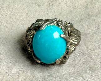 Sterling silver ring featuring a vibrant turquoise cabochon centerpiece. The band is ornately designed with detailed organic elements resembling leaves or vines. The turquoise stone is prominently set, securely held by prongs. The interior of the band shows slight wear, consistent with age and previous use. Comes in a size 6.