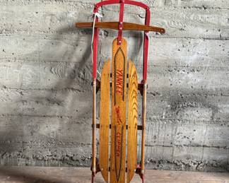 Wooden Flexible Flyer Yankee Clipper sled featuring steel runners painted in red and a wooden deck with printed branding. The sled includes a wooden crossbar for steering and a rope control. Signs of wear consistent with age and use, including scratches and minor rust on metal parts, are visible. A classic item for collectors or enthusiasts of vintage winter gear. Measures, 44” H x 20.5” W.