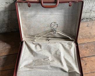 Brown leather suitcase adorned with various vintage travel decals from international destinations. Features brass hardware, interior cloth lining, and built-in hangers. Includes a monogram ‘L.M.’ on the leather exterior. Condition shows wear consistent with use, including scuffs and scratches on the leather and minor handling marks inside. No key.