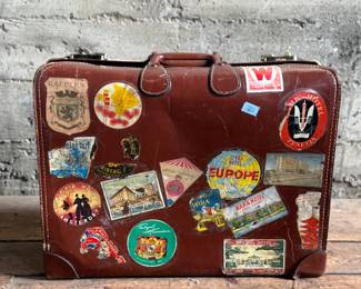 Brown leather suitcase adorned with various vintage travel decals from international destinations. Features brass hardware, interior cloth lining, and built-in hangers. Includes a monogram ‘L.M.’ on the leather exterior. Condition shows wear consistent with use, including scuffs and scratches on the leather and minor handling marks inside. No key.