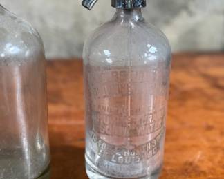 Vintage soda siphons featuring glass bodies and metal dispensers. One bottle has visible etched lettering, indicating it is from St. Louis, Missouri. Both show signs of wear and age, including discoloration, patina on the metal tops, and residue inside. These pieces are suitable as decorative or collectible items.