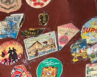 Brown leather suitcase adorned with various vintage travel decals from international destinations. Features brass hardware, interior cloth lining, and built-in hangers. Includes a monogram ‘L.M.’ on the leather exterior. Condition shows wear consistent with use, including scuffs and scratches on the leather and minor handling marks inside. No key.