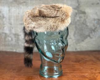 A fur cap featuring a raccoon tail, displayed on a handmade glass mannequin head. The cap’s material is natural fur, with a thick soft lining for comfort. The glass head is crafted from recycled glass and made in Spain. Both the cap and glass head appear in good condition.