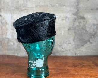 Black faux fur hat with a circular design, displayed on a translucent green glass head made from recycled materials and handcrafted in Spain. The interior of the hat is lined with fabric and features small red details. The glass head has a textured surface with a visible sticker stating its recycled origin.