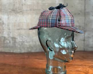 Tartan deerstalker cap featuring a classic red, blue, and white plaid pattern with a black ribbon detail on top. Inner lining crafted from black fabric for added comfort, and a label marked “Tartan Gift Shop by Lynton” is present. Displayed on a handblown glass head form, made from recycled glass in Spain, as indicated by the sticker. A distinctive decorative or collectible piece.