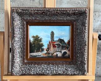 Framed oil painting depicting a European village scene featuring a bridge, architecture, and figures in a cobblestone street. The artwork comes in an ornately carved frame with intricate detailing. The reverse side is covered with paper backing and equipped with a hanging wire. Signed lower right by artist and measures in frame, 16” H x 18” W.
