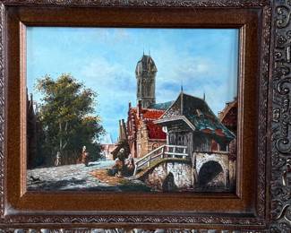 Framed oil painting depicting a European village scene featuring a bridge, architecture, and figures in a cobblestone street. The artwork comes in an ornately carved frame with intricate detailing. The reverse side is covered with paper backing and equipped with a hanging wire. Signed lower right by artist and measures in frame, 16” H x 18” W.