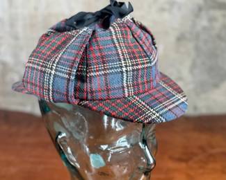 Tartan deerstalker cap featuring a classic red, blue, and white plaid pattern with a black ribbon detail on top. Inner lining crafted from black fabric for added comfort, and a label marked “Tartan Gift Shop by Lynton” is present. Displayed on a handblown glass head form, made from recycled glass in Spain, as indicated by the sticker. A distinctive decorative or collectible piece.