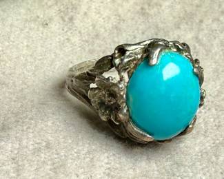 Sterling silver ring featuring a vibrant turquoise cabochon centerpiece. The band is ornately designed with detailed organic elements resembling leaves or vines. The turquoise stone is prominently set, securely held by prongs. The interior of the band shows slight wear, consistent with age and previous use. Comes in a size 6.