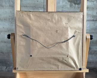 Framed oil painting depicting a European village scene featuring a bridge, architecture, and figures in a cobblestone street. The artwork comes in an ornately carved frame with intricate detailing. The reverse side is covered with paper backing and equipped with a hanging wire. Signed lower right by artist and measures in frame, 16” H x 18” W.