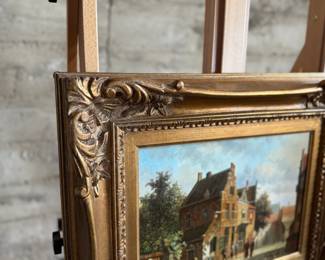 Framed oil on panel painting depicting a detailed village scene with several figures and buildings. Signed by the artist in the lower right corner. Encased in an ornate, gilt wooden frame with floral details. Measures in frame, 20.75” H x 24.75” W.
