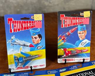 Two Matchbox Thunderbirds action figures (Scott Tracy and Alan Tracy) on original carded packaging, and two Micro Machines National Geographic Collections: #3 Moon Explorers and #4 Space Pioneers. All items are in original unopened packaging with visible wear consistent with age. Educational and collectible sets representing iconic figures and space exploration themes.