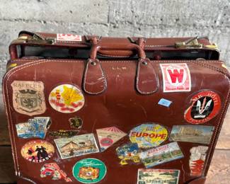 Brown leather suitcase adorned with various vintage travel decals from international destinations. Features brass hardware, interior cloth lining, and built-in hangers. Includes a monogram ‘L.M.’ on the leather exterior. Condition shows wear consistent with use, including scuffs and scratches on the leather and minor handling marks inside. No key.