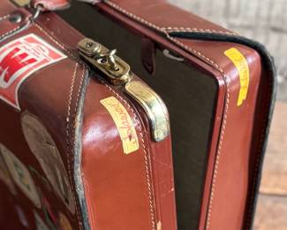 Brown leather suitcase adorned with various vintage travel decals from international destinations. Features brass hardware, interior cloth lining, and built-in hangers. Includes a monogram ‘L.M.’ on the leather exterior. Condition shows wear consistent with use, including scuffs and scratches on the leather and minor handling marks inside. No key.