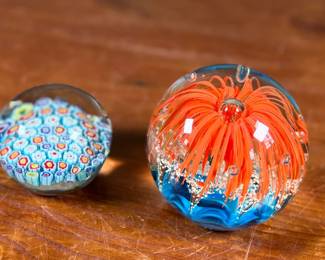 Three glass paperweights featuring intricate floral and abstract designs. Includes one large piece with orange floral motifs and two smaller pieces showcasing millefiori patterns in vibrant colors. These paperweights are in excellent condition with no visible chips or cracks. No visible signatures seen.