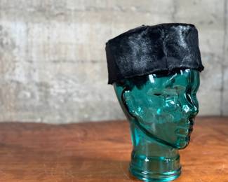 Black faux fur hat with a circular design, displayed on a translucent green glass head made from recycled materials and handcrafted in Spain. The interior of the hat is lined with fabric and features small red details. The glass head has a textured surface with a visible sticker stating its recycled origin.