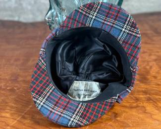 Tartan deerstalker cap featuring a classic red, blue, and white plaid pattern with a black ribbon detail on top. Inner lining crafted from black fabric for added comfort, and a label marked “Tartan Gift Shop by Lynton” is present. Displayed on a handblown glass head form, made from recycled glass in Spain, as indicated by the sticker. A distinctive decorative or collectible piece.