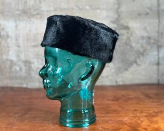 Black faux fur hat with a circular design, displayed on a translucent green glass head made from recycled materials and handcrafted in Spain. The interior of the hat is lined with fabric and features small red details. The glass head has a textured surface with a visible sticker stating its recycled origin.